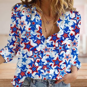 May include: A white, long-sleeved shirt with a vibrant pattern of red and blue stars. The stars vary in size and design, creating a patriotic and eye-catching look. The shirt has a V-neck and button details.
