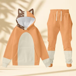 May include: An orange and white fox-themed tracksuit. The hooded top has a fox face design on the hood, a white belly, and a front pocket. The matching joggers have a drawstring waist and white cuffs. The outfit is made of a soft material.