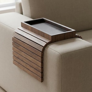 Puede incluir: Una bandeja de brazo de sofá de madera con acabado marrón oscuro. La bandeja tiene una parte superior rectangular y un panel lateral de listones que se curva para adaptarse al brazo del sofá. La bandeja está diseñada para sostener bebidas, aperitivos u otros artículos.