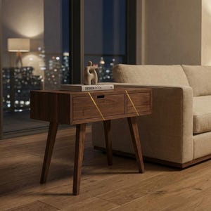 May include: A dark wood nightstand with four tapered legs and a single drawer featuring a horizontal cutout handle. The drawer front is accented with two thin, diagonal gold-colored lines. A book titled DESIGN BOOK sits on top.