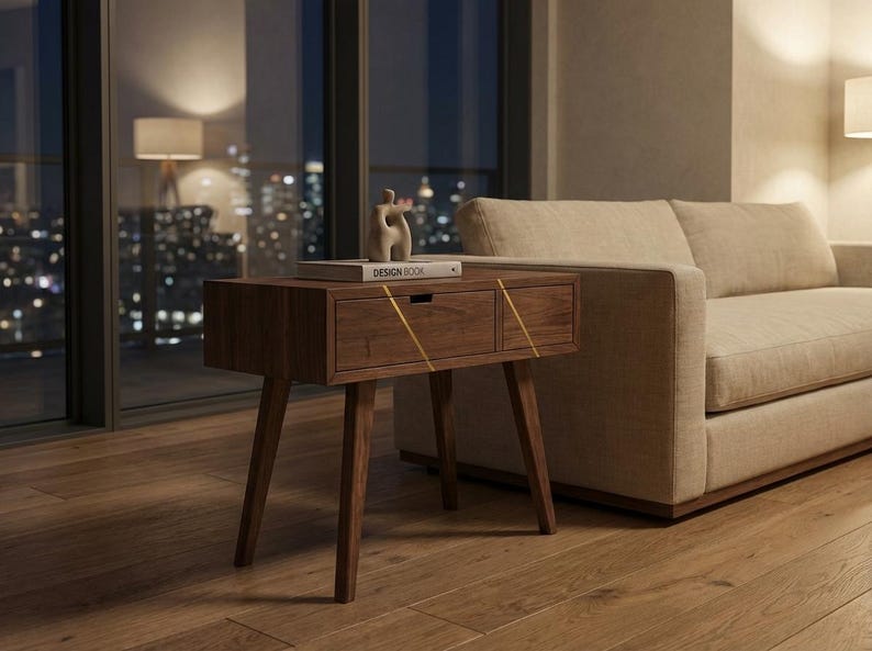 May include: A dark wood nightstand with four tapered legs and a single drawer featuring a horizontal cutout handle. The drawer front is accented with two thin, diagonal gold-colored lines. A book titled DESIGN BOOK sits on top.