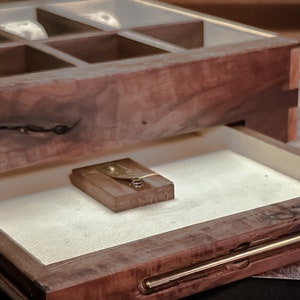 May include: A wooden box with a drawer that opens to reveal a small, rectangular object. The box has a dark brown finish and a natural wood grain. The drawer is lined with a white fabric.