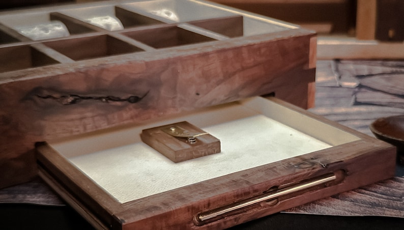 May include: A wooden box with a drawer that opens to reveal a small, rectangular object. The box has a dark brown finish and a natural wood grain. The drawer is lined with a white fabric.