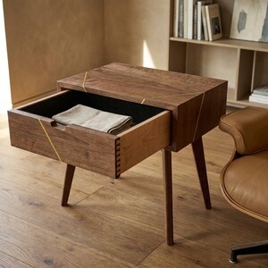 May include: A dark wood nightstand with four tapered legs and a single pull-out drawer. The drawer front features a light-colored wood side with visible dovetail joinery and thin gold-toned inlay lines across the top and front surface.