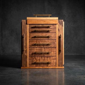 May include: A wooden jewelry chest featuring a natural wood grain finish with five horizontal drawers and two side doors. Each drawer and door is equipped with a textured, horizontal brass-toned metal handle. The top surface includes a small handle.