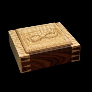 Könnte beinhalten: Eine Holzkiste mit einem Deckel aus hellem Holz und einem Sockel aus dunklem Holz. Der Deckel hat ein geschnitztes Unendlichkeitszeichen. Die Kiste hat eine Gehrungskante.