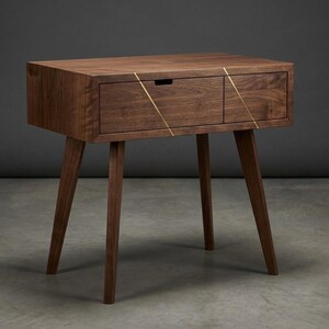 May include: A dark brown wooden nightstand featuring four tapered legs and a single drawer. The drawer front is accented with two thin, diagonal gold-colored metal inlays that create a geometric pattern across the wood grain surface.