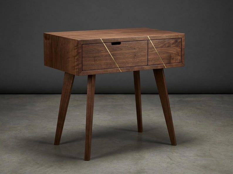 May include: A dark brown wooden nightstand featuring four tapered legs and a single drawer. The drawer front is accented with two thin, diagonal gold-colored metal inlays that create a geometric pattern across the wood grain surface.