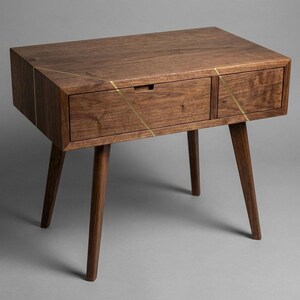 May include: A mid-century modern style wooden nightstand with a drawer and a cabinet. The nightstand is made of dark brown wood with brass accents and has four tapered legs. The drawer has a small, rectangular handle.