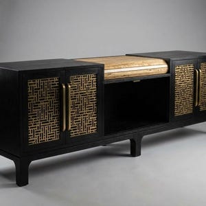 May include: A black wooden cabinet with gold-colored geometric patterned doors and handles. The cabinet has a light-colored wooden top and an open shelf. The cabinet is a modern piece of furniture.