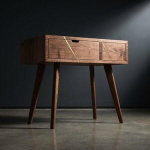 May include: A mid-century modern wooden side table with a drawer and angled legs. The table is made of dark wood and features brass inlay accents. The design is minimalist and the table is suitable for a living room or bedroom.