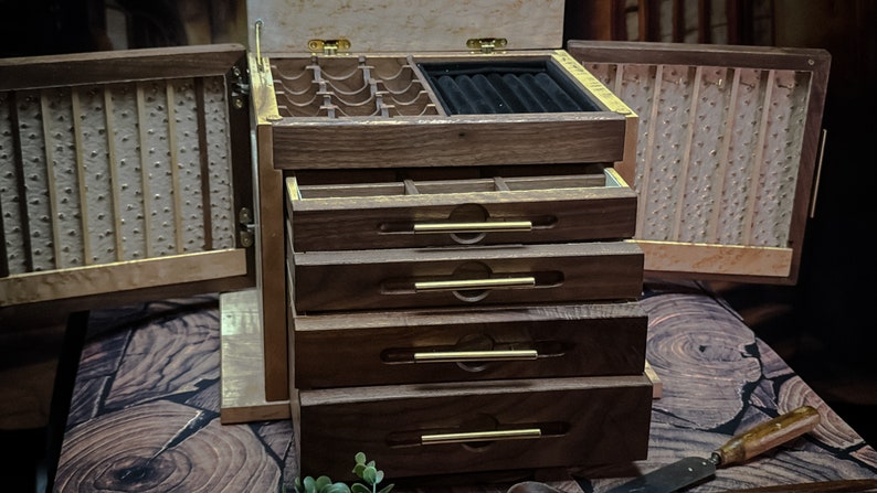 May include: A wooden jewelry box with a hinged lid and multiple drawers. The box is made of dark wood with brass accents. The lid is open and reveals a compartment with a black velvet lining. The box has four drawers with brass handles.