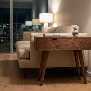 May include: A mid-century modern style wooden side table with two drawers and angled legs. The table is a dark brown color with gold accents. A book with the word "DESIGN" is on top, along with a small ceramic figurine.