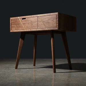 May include: A mid-century modern wooden nightstand with two drawers and angled legs. The nightstand is a warm brown color with gold accent lines across the drawer fronts. The design is minimalist and stylish.