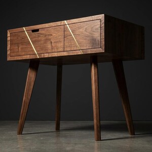 May include: A dark brown wooden side table featuring two drawers with a visible grain pattern. The front of the drawers is accented by two thin, diagonal gold-colored metal inlays that create a geometric pattern across the drawer faces.