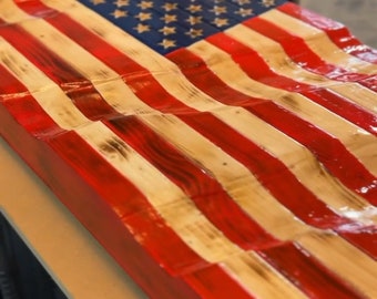May include: A wooden American flag with red, white, and blue paint. The flag is made of individual wood planks that are arranged to create a wavy effect. The flag is painted in a distressed style, giving it a vintage look.