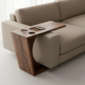 May include: A modern sofa with a built-in wooden side table. The table has a drink holder, a small compartment, and a slot for a remote. The sofa is upholstered in a neutral beige fabric, and the table is made of dark wood.