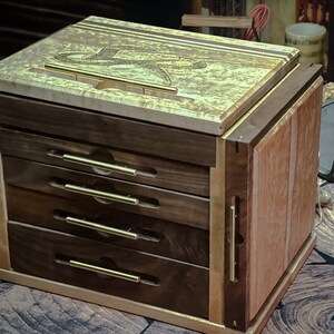 May include: A wooden box with four drawers and brass pulls. The top of the box is made of a light wood with a gold-colored finish. The box is sitting on a wooden surface.