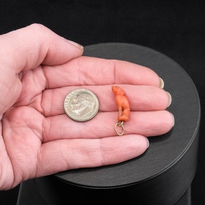 May include: A small, coral-colored figurine of a person, hanging from a gold chain. The figurine is about the size of a dime.