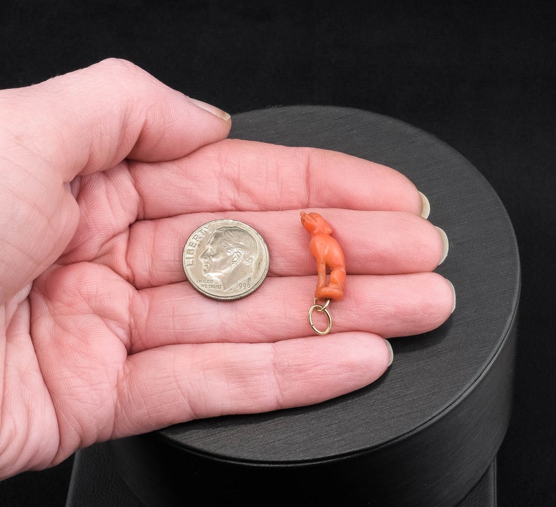 May include: A small, coral-colored figurine of a person, hanging from a gold chain. The figurine is about the size of a dime.