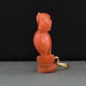 May include: A coral and gold charm in the shape of a sitting bird. The charm is a deep orange color and has a gold loop for attaching to a necklace or bracelet.