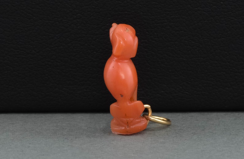 May include: A coral and gold charm in the shape of a sitting bird. The charm is a deep orange color and has a gold loop for attaching to a necklace or bracelet.