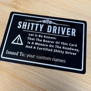 May include: A black rectangular card with white text that reads "SHITTY DRIVER." The card includes a warning symbol and the text "Let It Be Known: That The Bearer Of This Card Is A Menace On The Roadway, And A Certified Shitty Driver."