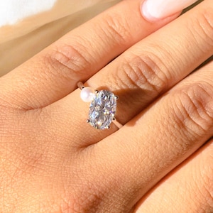 May include: A silver ring with a large, oval-cut diamond and a small pearl. The diamond reflects light. The ring is set on a delicate band. The ring is a piece of jewellery. The ring is a piece of jewellery.