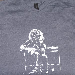 May include: Dark gray t-shirt featuring a white graphic of a musician playing a guitar. The design shows the musician seated, with a microphone and amplifier visible. The shirt is made of a soft material.