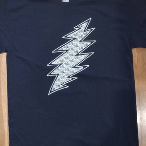 May include: Navy blue t-shirt featuring a white and blue lightning bolt design. The lightning bolt is filled with a repeating pattern of small, detailed images. The shirt is made of a soft material.