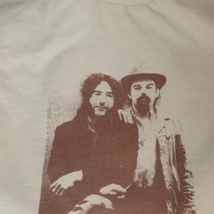 May include: A vintage white t-shirt with a brown print of two men. The men are wearing casual clothing and have a friendly expression. The man on the left has long hair and is wearing a jacket. The man on the right is wearing a hat and a jacket.