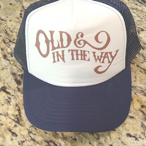 May include: A white and navy blue trucker hat with a brown embroidered design that says "Old & In The Way".