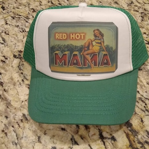 May include: Green and white trucker hat with a curved brim. The front panel features a vintage-style graphic with the words "RED HOT MAMA" and an image of a woman. The hat has a mesh back and a white button on top.