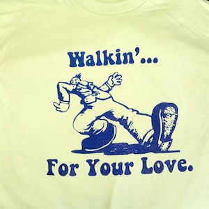 May include: A light yellow t-shirt with the words "Walkin'... For Your Love" in dark blue. The shirt features a blue cartoon illustration of a person walking. The shirt is made of a soft material.