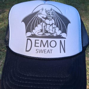 May include: A black and white trucker hat featuring a graphic of a winged demon figure on the front foam panel. Below the illustration, the text reads DEMON SWEAT in black capital letters. The hat has a black brim and black mesh side panels.