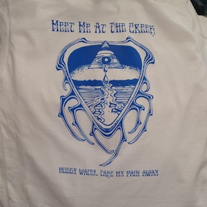 May include: White t-shirt with a blue graphic design. The design features an eye within a triangle, a water droplet, and the text "Meet Me At The Creek" and "Muddy Water, Take My Pain Away."