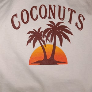 May include: A white t-shirt with a brown graphic of two palm trees and a setting sun. The text "COCONUTS" is printed in brown above the graphic.
