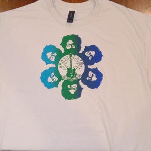 May include: White t-shirt featuring a colorful graphic design. The design includes six stylized faces in a flower-like arrangement, surrounding a guitar. The faces are in shades of blue and green.