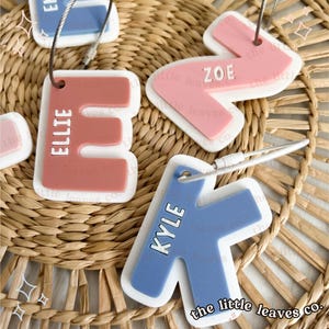 May include: Three personalised keychains with the names Ellie, Zoe, and Kyle. The keychains are made of silicone and have a white border. The keychains are in the shape of the first letter of each name. The Ellie keychain is pink, the Zoe keychain is pink, and the Kyle keychain is blue. The keychains are on a woven background.