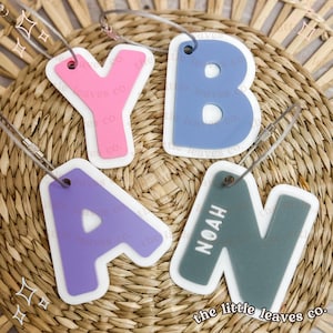 May include: Four colourful letter charms with white outlines, spelling out the name "YAN", with a fifth charm that says "NOAH" in white letters on a grey background. The charms are attached to silver rings and are displayed on a woven wicker surface.