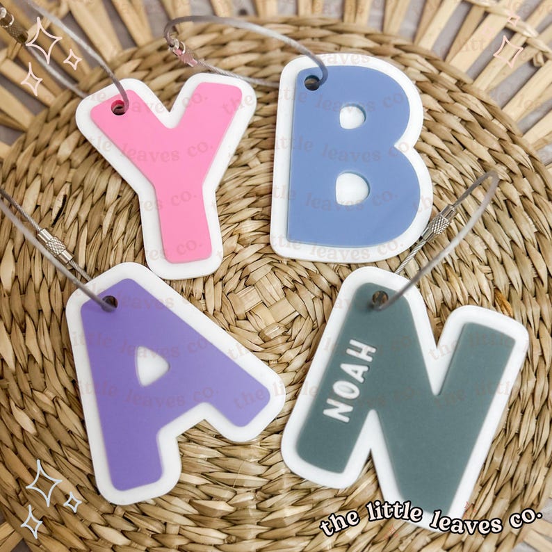 May include: Four colourful letter charms with white outlines, spelling out the name "YAN", with a fifth charm that says "NOAH" in white letters on a grey background. The charms are attached to silver rings and are displayed on a woven wicker surface.