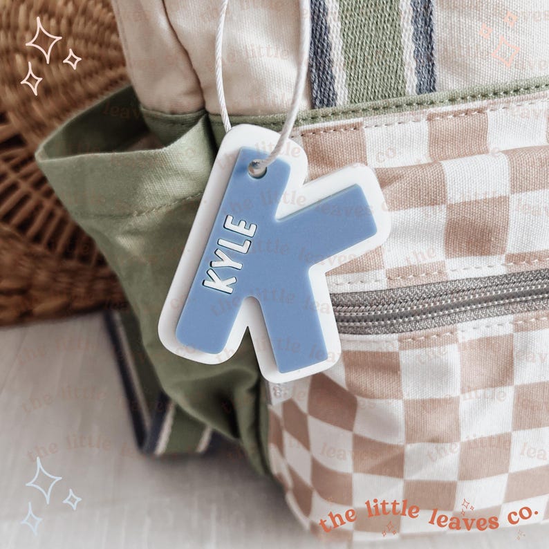 May include: A blue and white personalised name tag with the name "KYLE" in white lettering. The tag is shaped like a letter K and is attached to a white string.