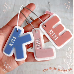 May include: Three personalised keychains with the names Kyle, Ellie and Leah. The keychains are made of white plastic with a blue, pink and red letter cutout. The letters are attached to a white plastic ring.