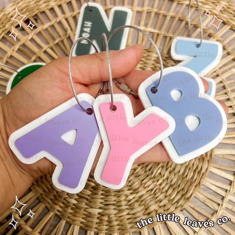 May include: A set of five colourful, personalised letter keychains. The letters are A, Y, B, N, and Z. The keychains are made of a soft, flexible material and have a white border. The keychains are attached to silver metal rings.