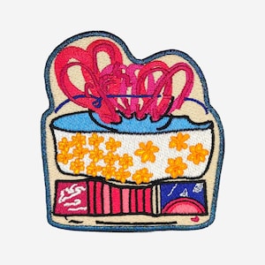 May include: Embroidered patch featuring a stylized design. The patch has a light blue border and a beige background. The design includes a blue bowl filled with yellow flowers and pink ribbon-like shapes.
