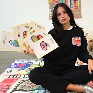 May include: A person holding a stack of art prints with yellow borders, each featuring colorful abstract illustrations. The person is wearing a black sweatshirt with a patch and black pants. The prints are labeled "ISABELLE GOUGENHEIM".