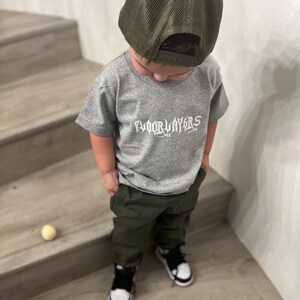 May include: A young person wearing a grey t-shirt with the text "FLOORLAYERS" in white, olive green pants, a green mesh trucker hat, and black and white sneakers.