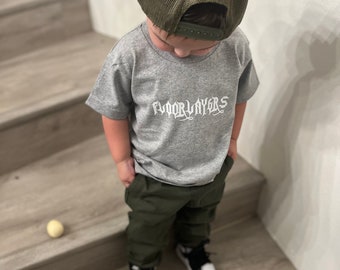 Può includere: Un giovane che indossa una maglietta grigia con il testo "FLOORLAYERS" in bianco, pantaloni verde oliva, un cappello da camionista in rete verde e scarpe da ginnastica bianche e nere.