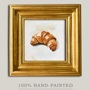 Puede incluir: Pintura de acuarela pintada a mano de un cruasán en un marco dorado. La pastelería es de color marrón dorado con pinceladas detalladas. El marco es de forma cuadrada con un borde decorativo. El texto en la parte inferior dice "100% HAND-PAINTED" y "ART BY LYNN".