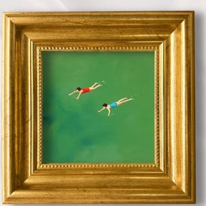 Puede incluir: Un cuadro cuadrado en un marco dorado que muestra a dos figuras nadando en agua verde. El texto "Lynn" y "100% Hand-Painted" aparece sobre el marco. La pintura representa a una figura con traje de baño rojo y otra con uno azul.